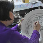 khatoun hand painting a large bowl - Balian - Armenian Ceramics A woman with short brown hair and glasses, wearing a purple sweater, is hand-painting an intricate design of birds and flowers on a large white ceramic plate in the workshop of Balian Pottery of Jerusalem.