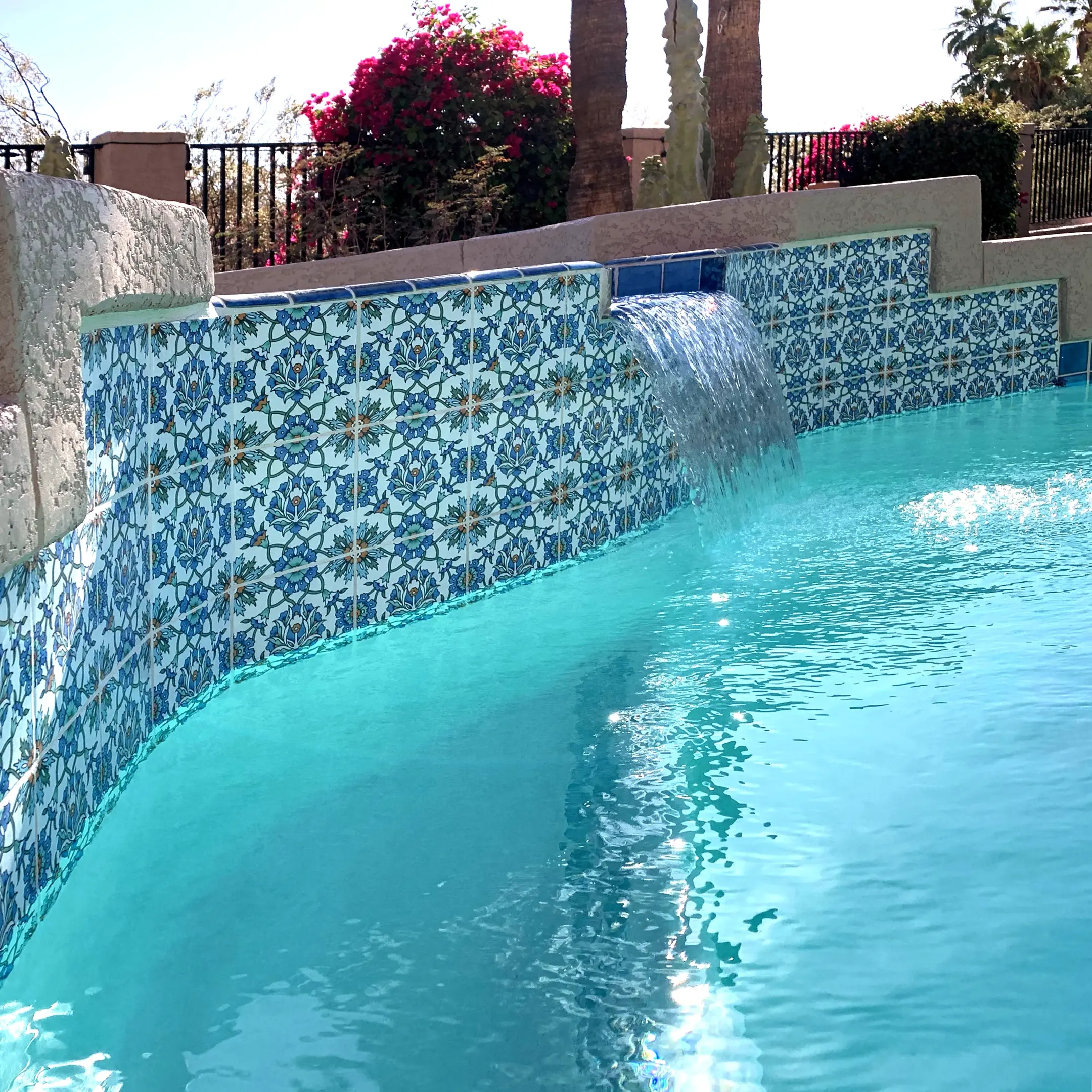The image features a beautifully curved swimming pool adorned with 6x6 inch porcelain tiles that reflect sunlight, situated next to a raised spa wall. The pool design incorporates stylish Balian porcelain waterline tiles, enhancing the luxurious feel of the outdoor area.