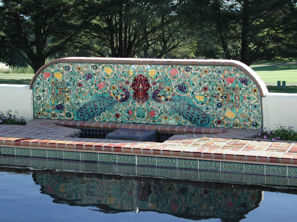 Hand-painted ceramic tile mural on an outdoor fountain wall beside a swimming pool, featuring peacock, floral and arabesque designs in teal and red